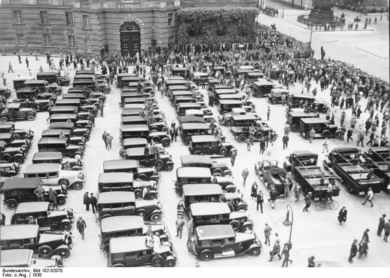 Rows of black motor cars parked in a large square, with crowds of pedestrians and a neoclassical building behind.