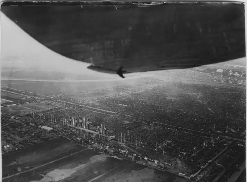 Aerial view of a huge field densely filled with people in blocks, with stands and flagpoles