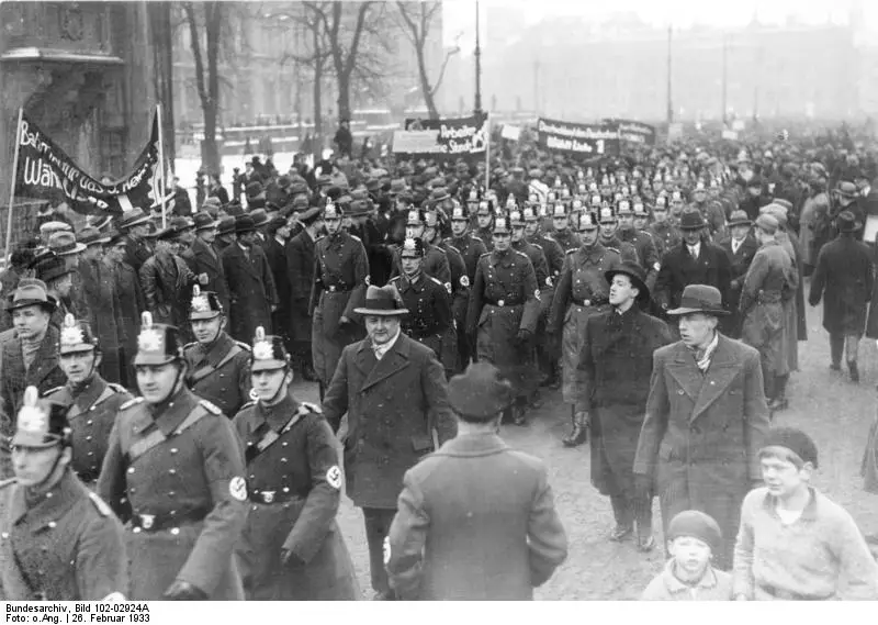 Columns of police in shakos and greatcoats, swastika armbands on their sleeves, march among men in hats and overcoats; banners are carried behind them.