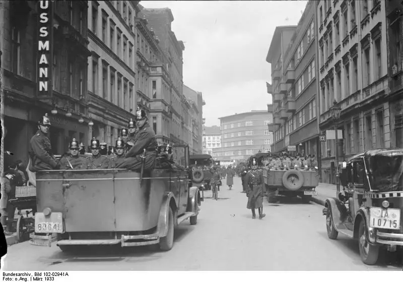 A street of tall tenement buildings with open police cars full of helmeted officers parked along it and a few figures on the pavements.
