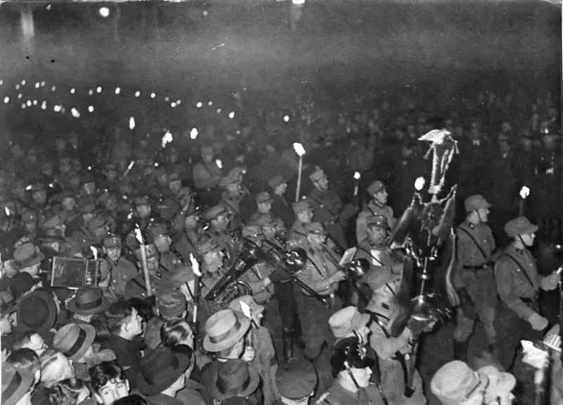At night, uniformed men carrying torches and brass instruments march through a dense crowd; a tall band standard hung with horsetails rises above them