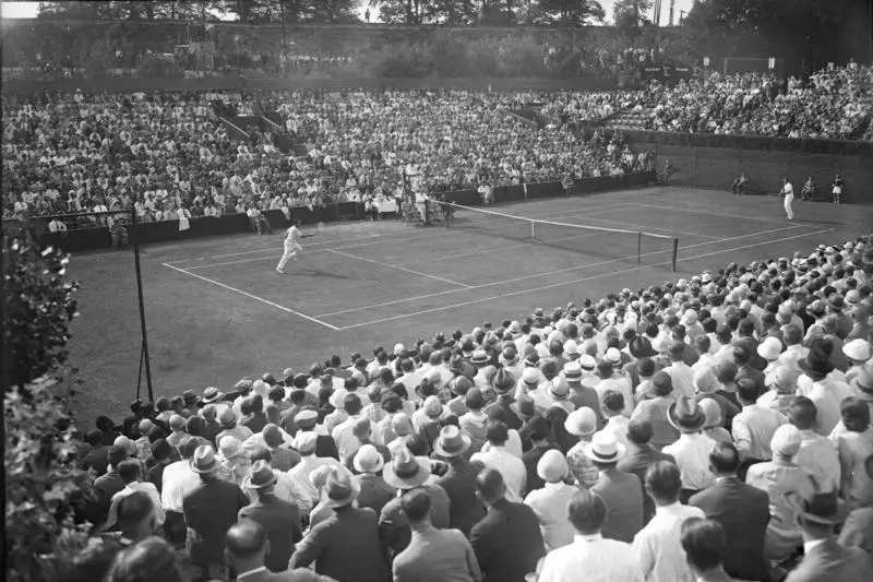 A tennis court seen from above with two players and huge crowds packed in stands and on terraces around it