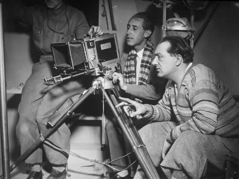 Three men crouch around a film camera on a tripod in a studio; the man at right, in a patterned pullover, points past the lens.