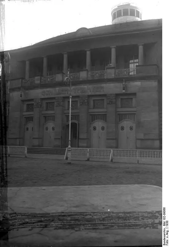 The stone facade of a theatre with a colonnaded upper storey and a row of arched doors behind a low fence, the name 'Schiller-Theater' across the front.