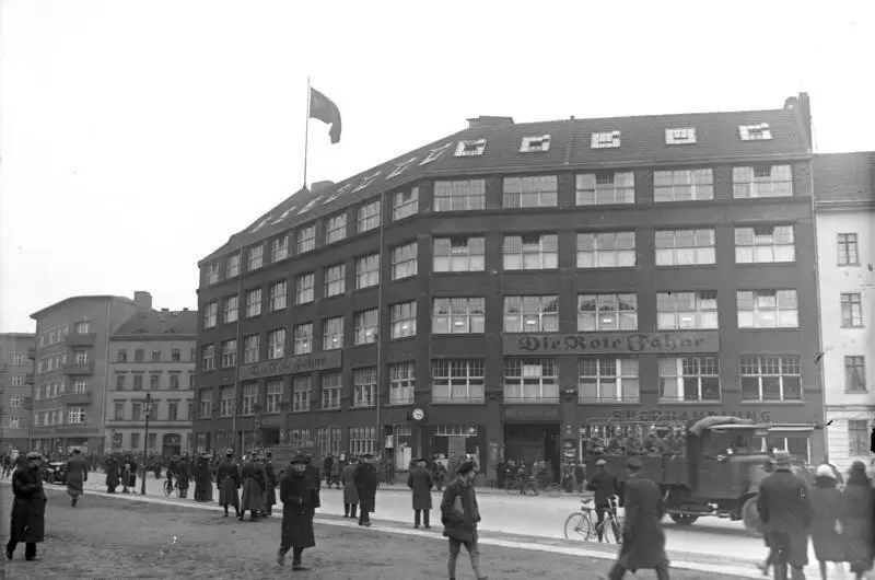 A large five-storey office building with a flag on the roof and the lettering "Die Rote Fahne", with police and onlookers in the open square in front.