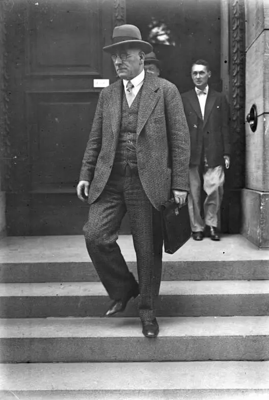 An older man in a hat and tweed suit carrying a briefcase walks down stone steps