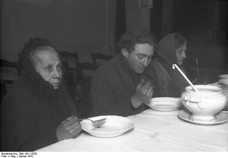An elderly woman in a headscarf, a young man and another woman eat soup at a table with a white cloth and a large tureen.