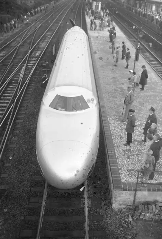 A long, low, streamlined railcar with a large propeller at its rear standing on tracks at a railway station.