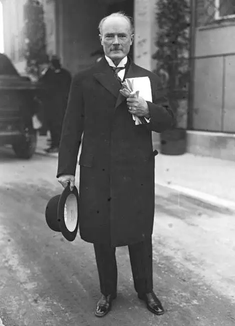 A tall elderly man in a dark overcoat holds a bowler hat and papers in front of a building entrance.