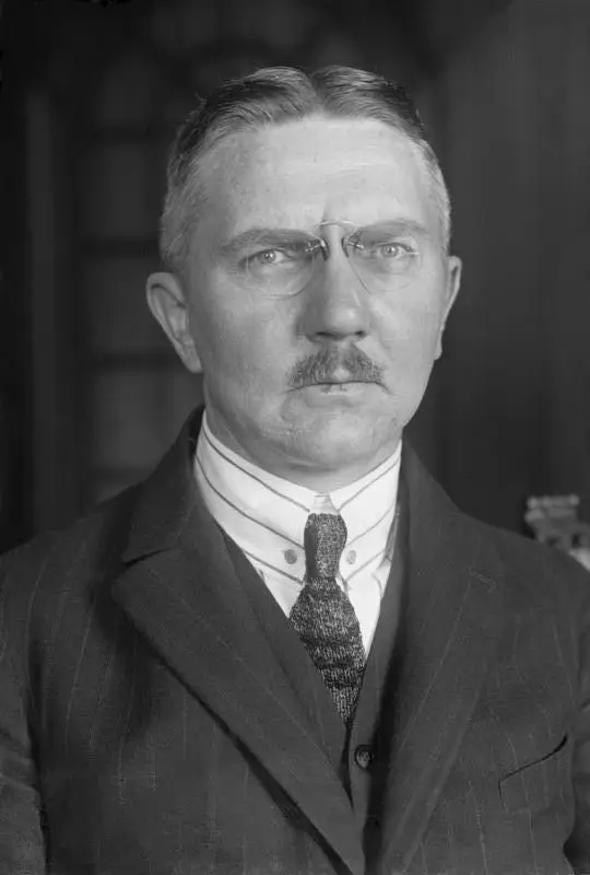 Portrait of a man with close-cropped hair, a moustache and pince-nez, wearing a striped collar and dark suit.