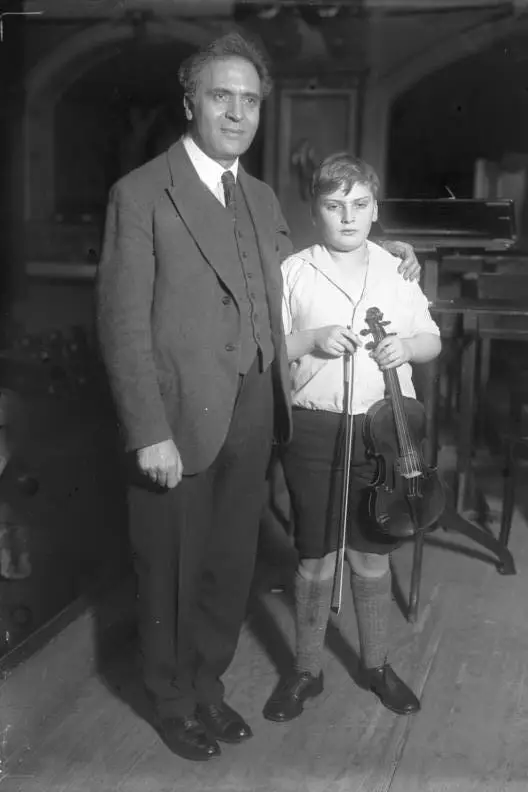 A middle-aged man in a suit with his hand on the shoulder of a boy holding a violin and bow.