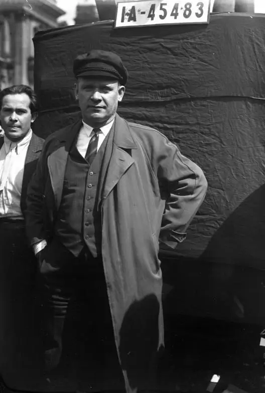 Ernst Thälmann in a peaked cap, tie and long open coat, standing in front of a covered vehicle with the registration plate IA-45483; another man stands behind him at left.