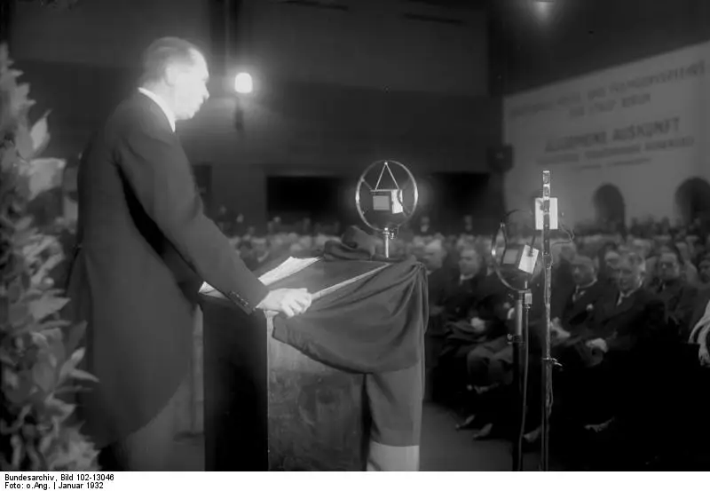 A tall man in a dark suit speaks at a draped lectern with two microphones before a seated audience in a hall.
