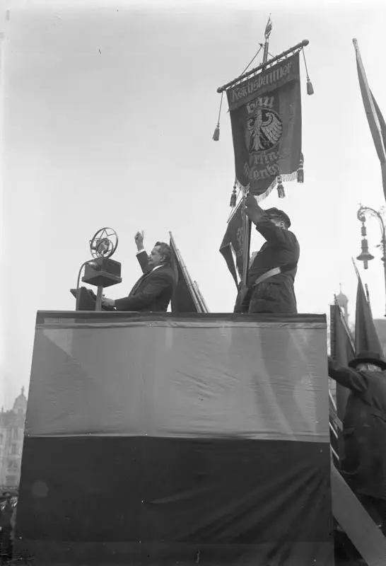 A man at a draped rostrum with a microphone raises his arm; beside him a man in uniform holds a banner with an eagle.