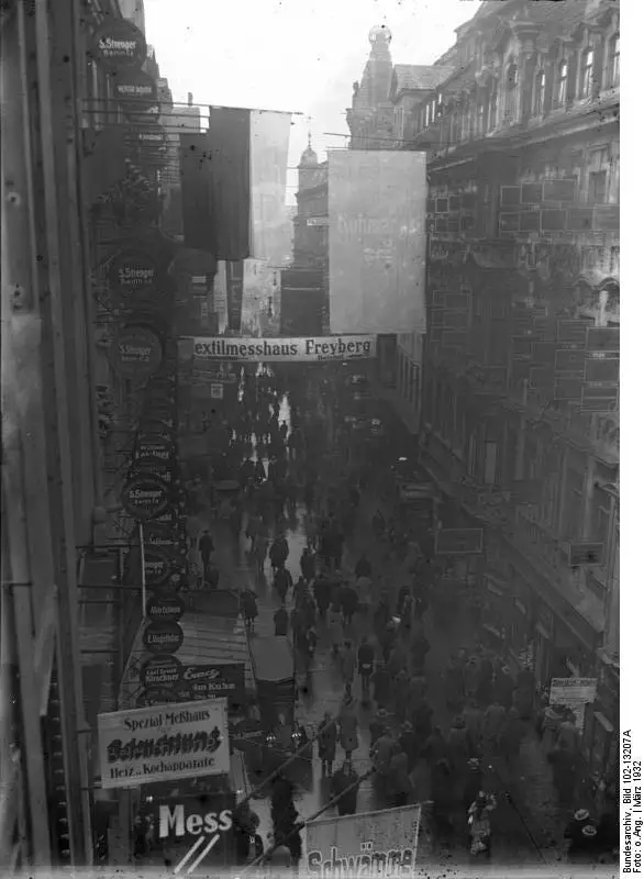 A narrow city street seen from above, crowded with pedestrians between tall buildings hung with signs and banners.