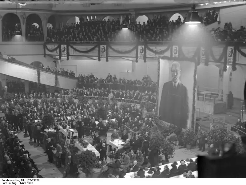 Interior of a vast hall with tiered balconies and a crowd seated at tables on the floor, with a huge portrait of Hindenburg hanging at the far end.