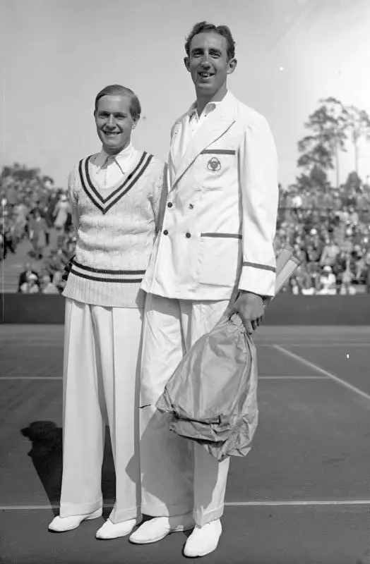 Two tennis players in white flannels stand side by side holding rackets.
