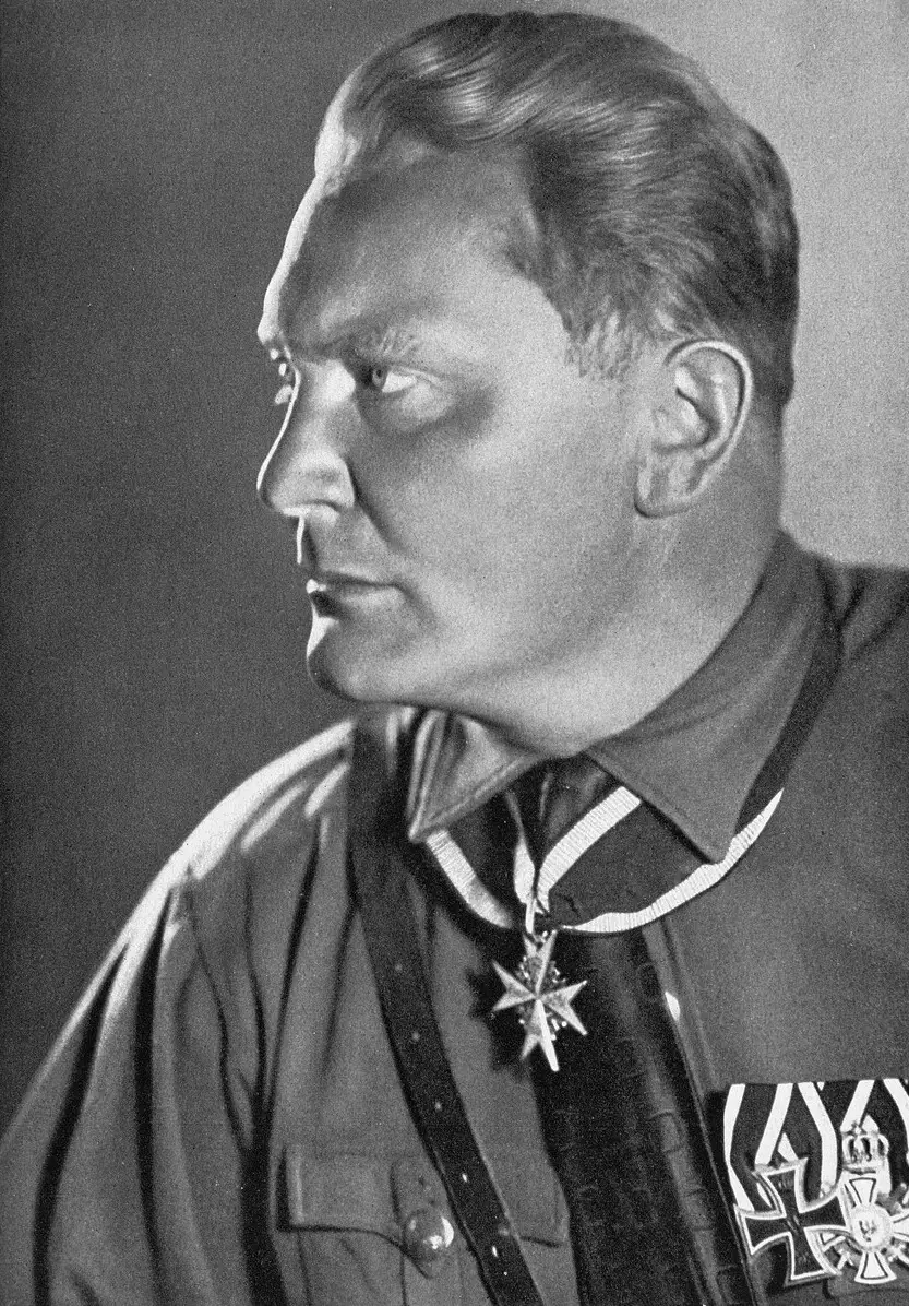 Profile portrait of a heavy-set man in a brown shirt with a cross-belt, wearing a cross on a ribbon at his neck and medals on his chest.