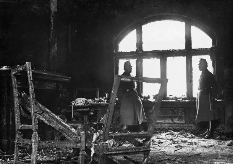 Interior of a fire-gutted room with charred chair frames in the foreground and two uniformed policemen standing by a large arched window.