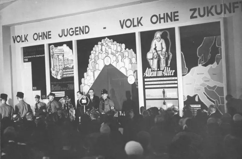 A speaker at a lectern on a stage flanked by uniformed men, beneath a large banner and population-policy posters, with an audience in the foreground.