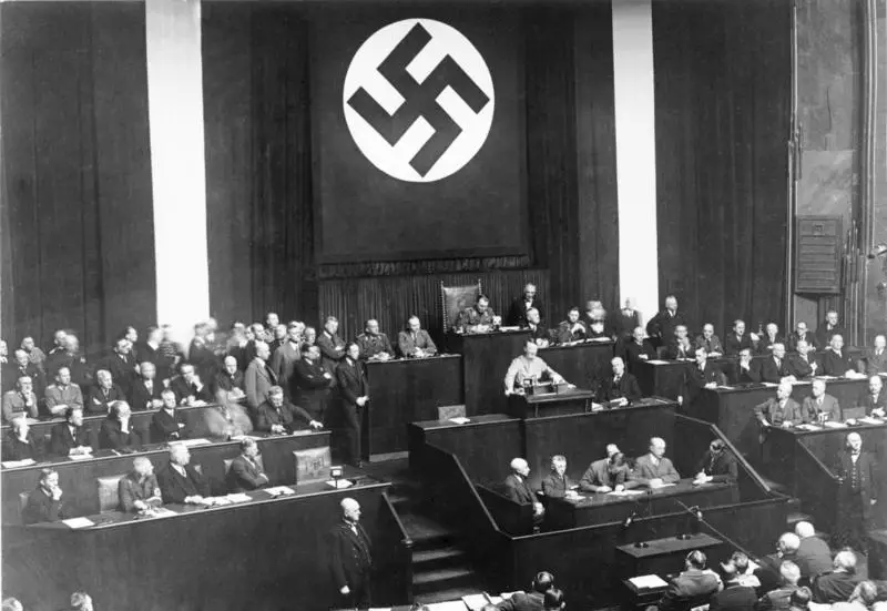 A theatre converted into a parliament: a speaker at a central rostrum, tiers of seated men around him and a huge swastika banner on the wall behind.