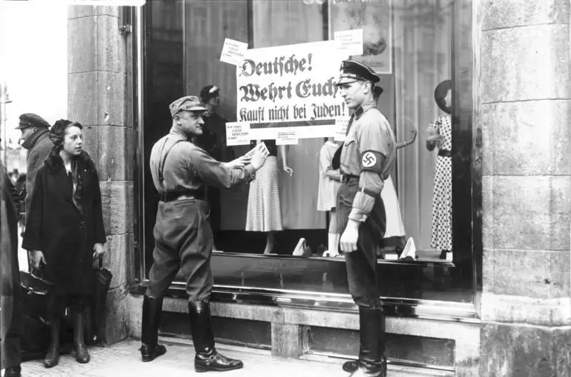 Two uniformed SA men at a large shop window displaying dresses; one presses a printed placard against the glass while a young woman with a handbag and other passers-by look on.