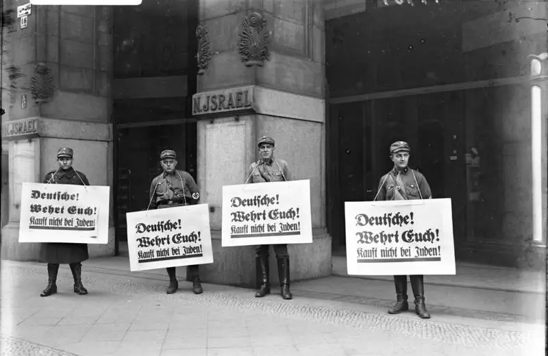 Four uniformed SA men stand in a row in front of a stone shopfront marked N. ISRAEL, each wearing a large placard reading 'Deutsche! Wehrt Euch! Kauft nicht bei Juden!'.