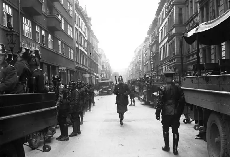 A narrow street between tall buildings filled with police lorries; uniformed men in shakos and helmets line both sides while an officer walks down the middle.