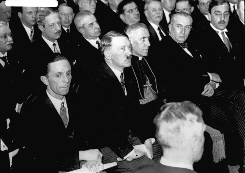 Rows of seated men in dark suits; in front, Goebbels, Hitler and a cleric in vestments with a pectoral cross listen to a speaker.