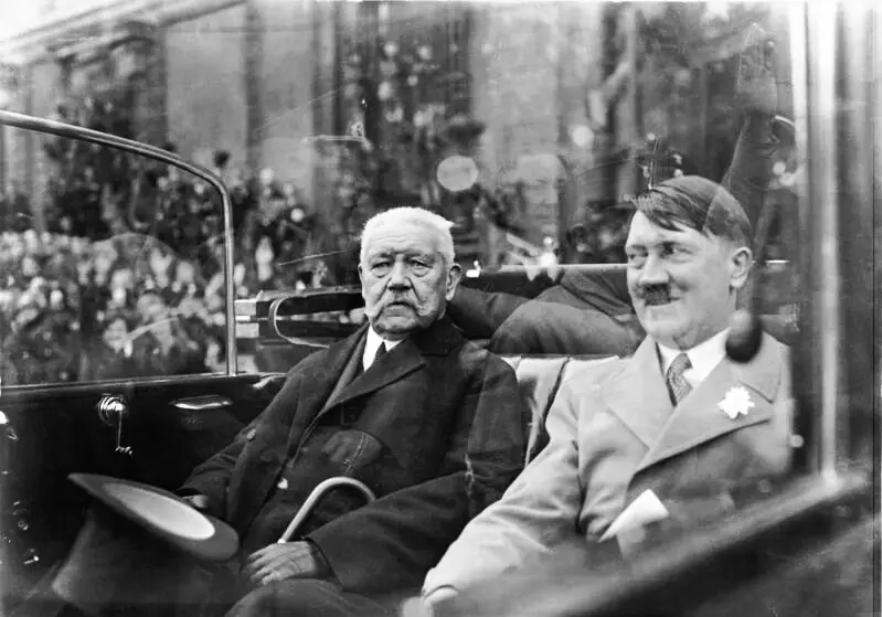An open car with two men in the back seat passing lines of saluting uniformed men