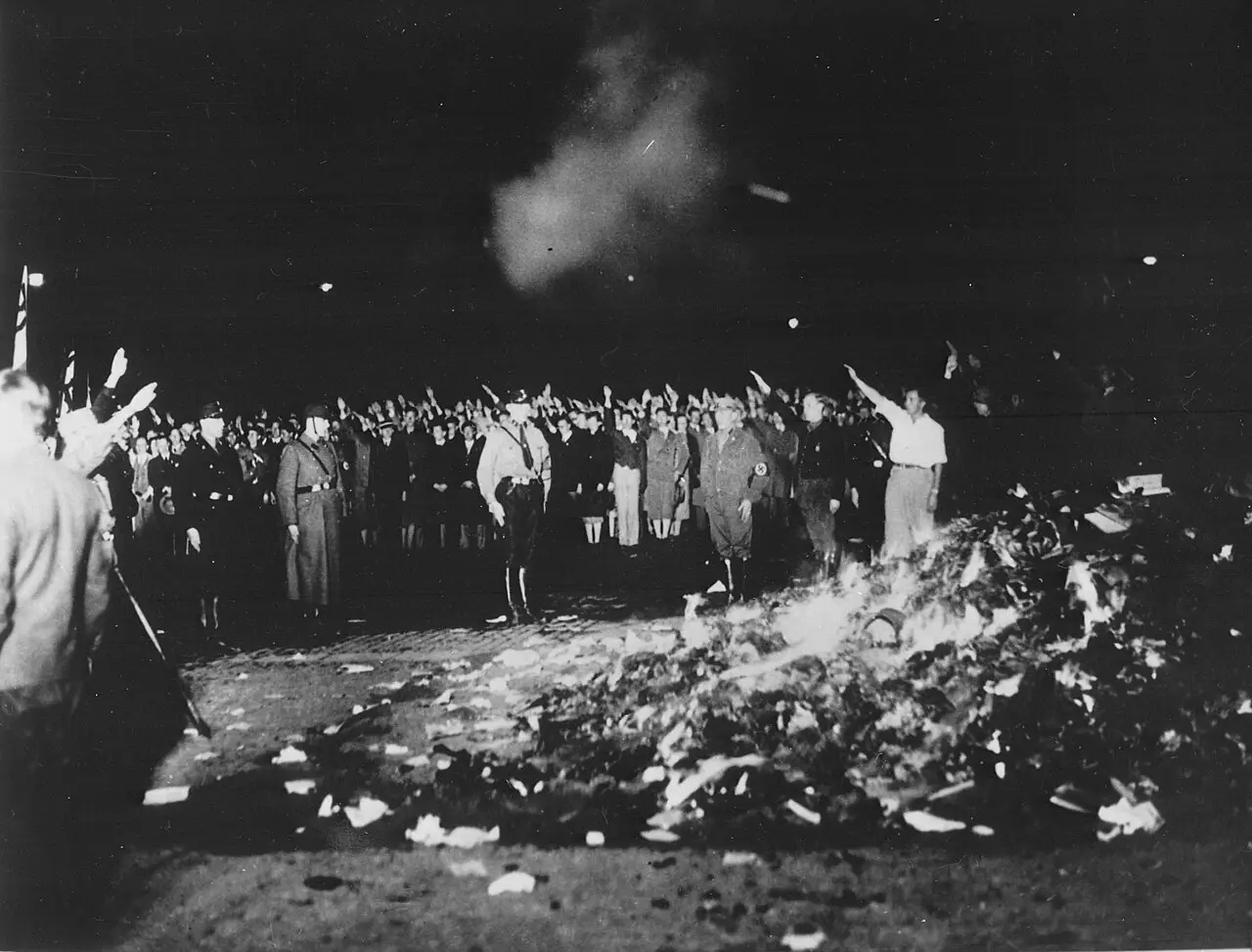 A large heap of burning books and papers at night, with uniformed men and a crowd behind it raising their arms in salute