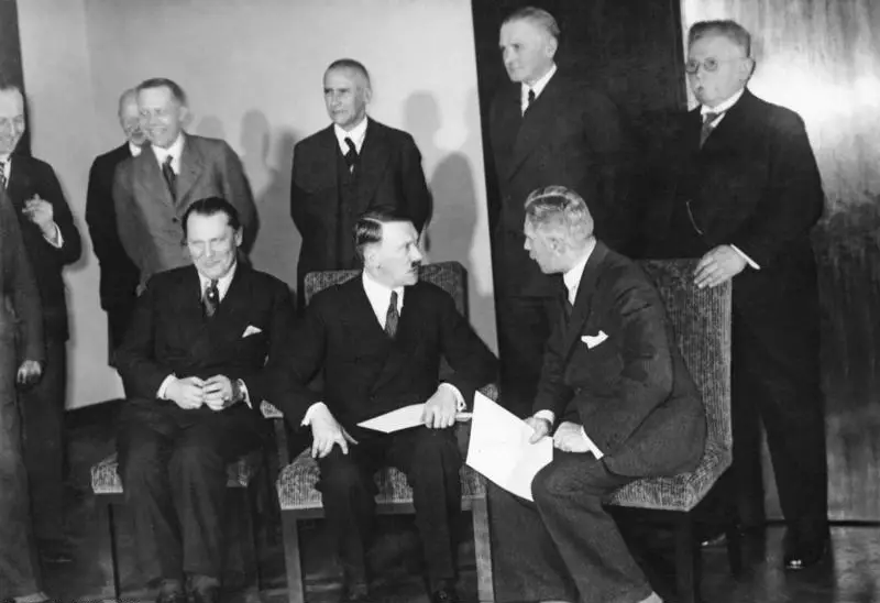 Seven men in dark suits standing in two rows in an office