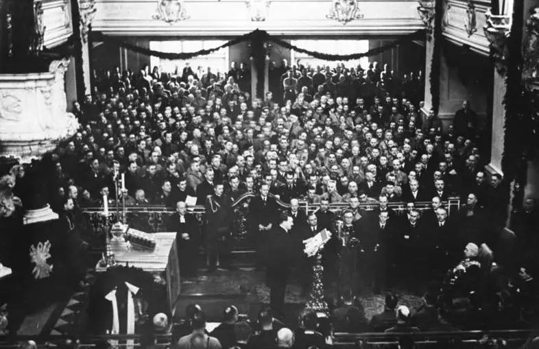 A crowded baroque church interior: rows of people in dark clothes, a speaker at a lectern and an elderly man seated in an armchair.