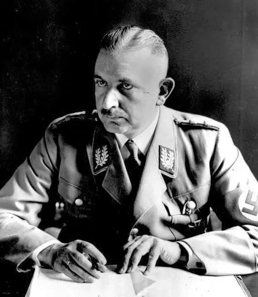 Portrait of a man with a moustache and swept-back hair in a light party uniform with an armband, seated at a desk with a pen in his hand.