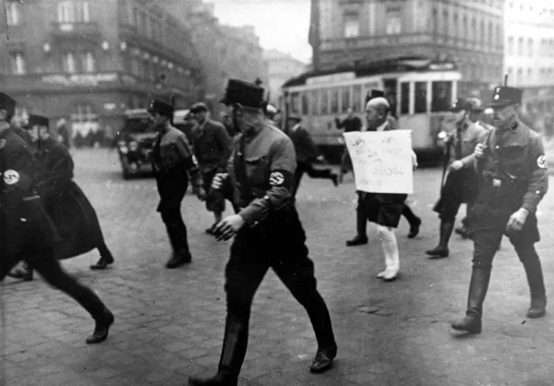 A man in a dark jacket and trousers cut off below the knee walks barefoot across a square with a board hanging from his neck, flanked by uniformed men.