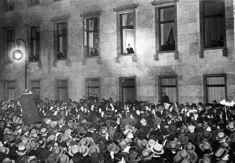 A crowd in hats below a lamp-lit stone façade; a man stands at an open first-floor window