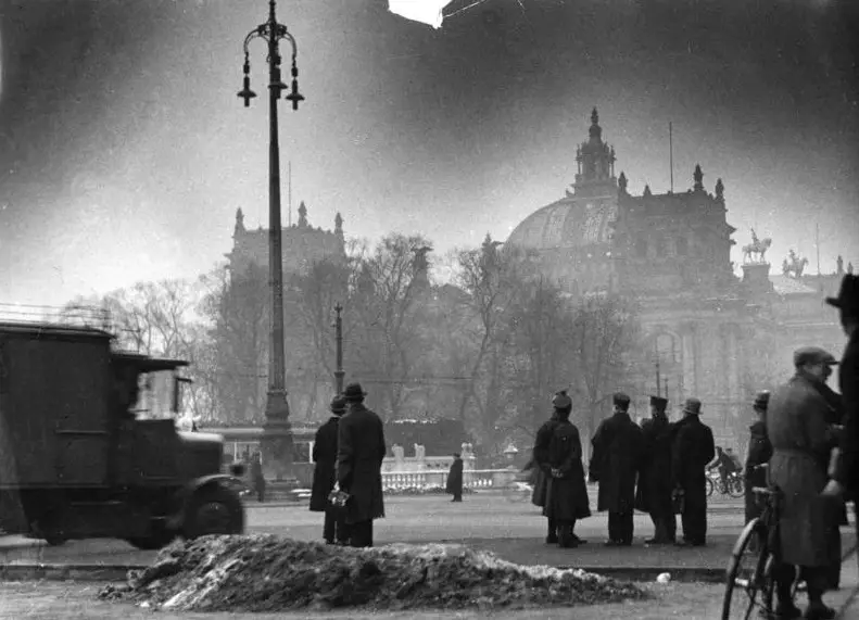 Street scene with a lorry, a lamp post and groups of men in coats looking towards the Reichstag building in the background.