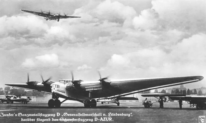 A huge four-engined monoplane parked on a field while a second large aircraft flies above it