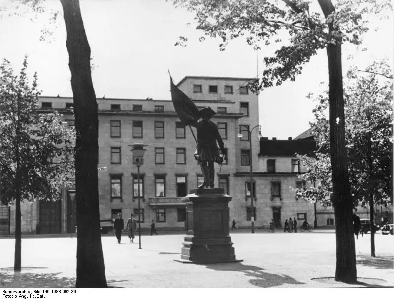 A plain four-storey office building behind trees and a bronze statue of an officer holding a flag on a tall plinth