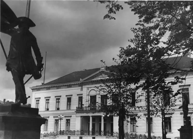 A two-storey classical palace with a balcony, seen past trees and the dark silhouette of a statue of an eighteenth-century soldier.