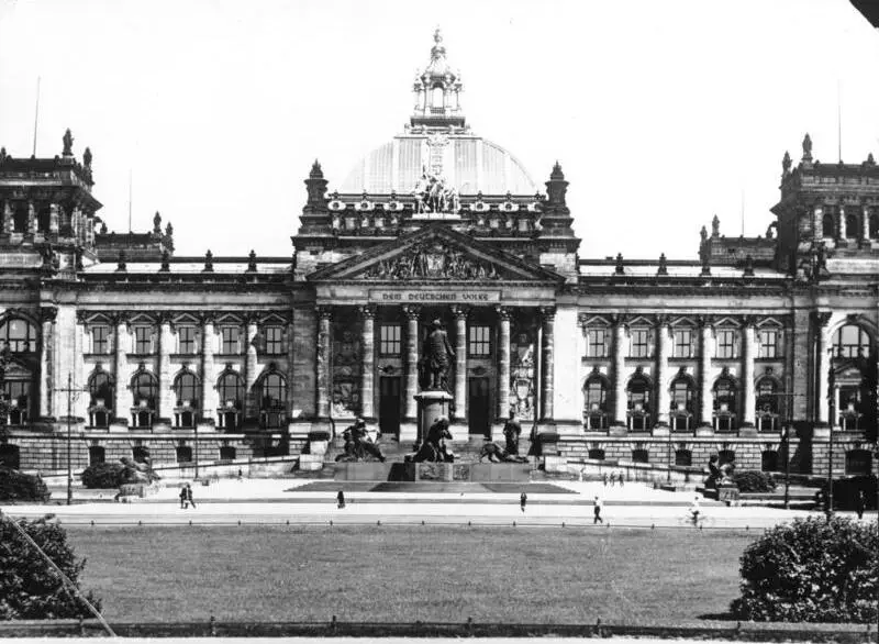 A large stone parliament building with a glass and steel dome and a columned portico, a monument on a plinth in front, seen across a lawn