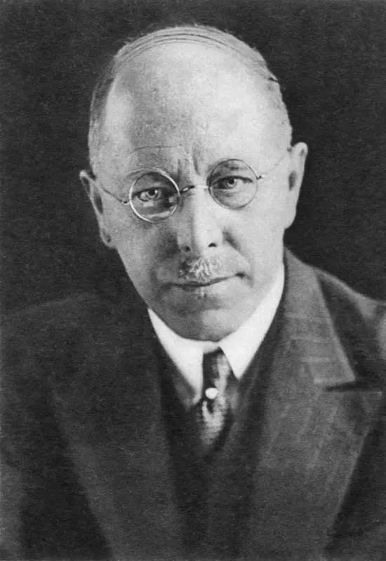 Head-and-shoulders portrait of a bald man with a moustache and spectacles in a dark suit.
