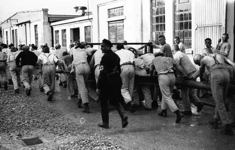 A group of men in work clothes pull a heavy wooden wagon across a yard while a guard in a dark uniform stands in front of them.