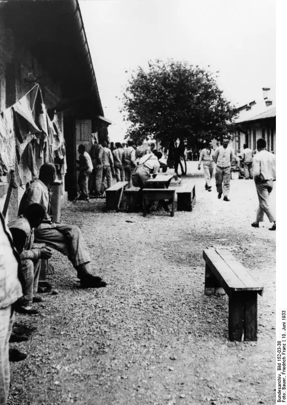 Men in work clothes sitting on benches and walking on a gravel yard between long low barrack buildings, with laundry hanging under the eaves.