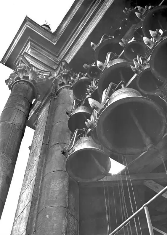 Rows of bronze bells hung in a frame inside a church tower, seen from below beside a carved stone column.