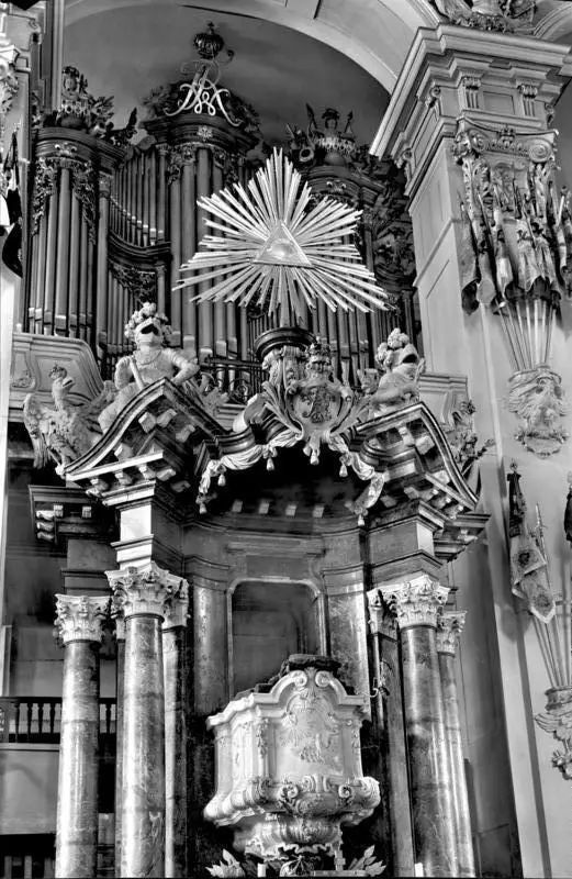 Ornate baroque church interior with a marble pulpit between columns, a radiant sunburst above it and organ pipes behind.
