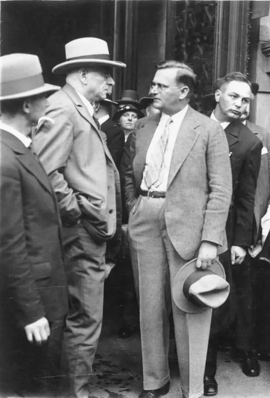 Two men talking outside a stone doorway, one holding his hat, with onlookers behind them