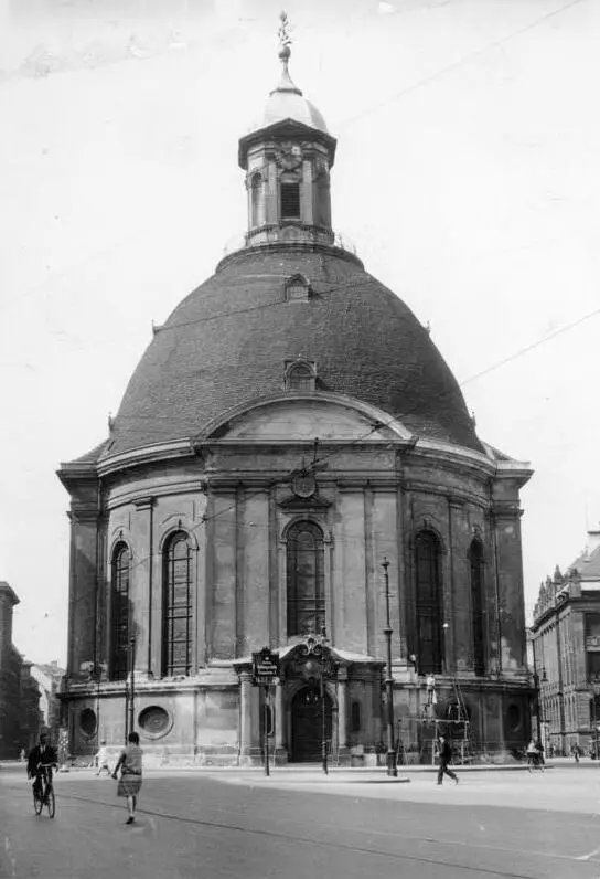 A domed baroque church with a lantern on top, standing at a street corner; a cyclist and a few pedestrians pass in the foreground.