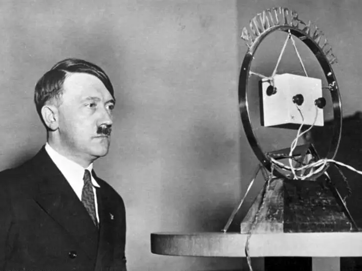 A man in a dark suit and tie stands beside a large ring-mounted microphone