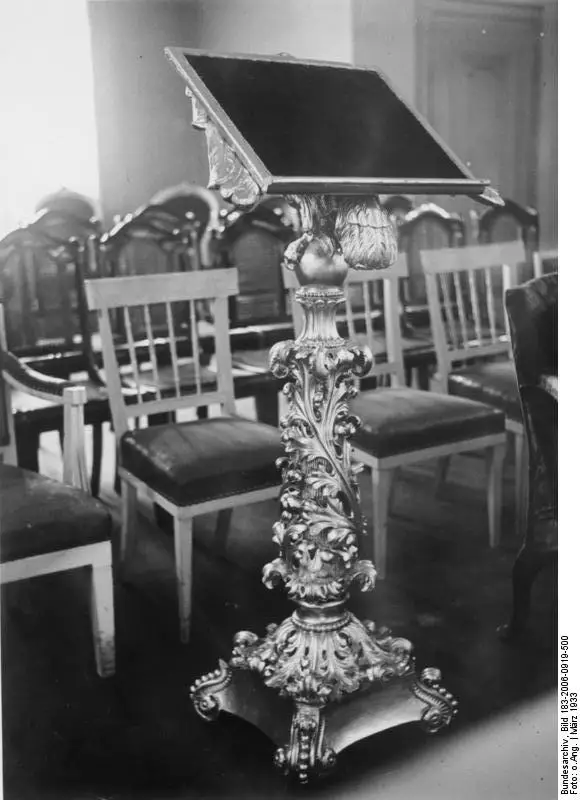 An ornate gilded lectern on a carved stand in front of rows of upholstered chairs in a church interior.